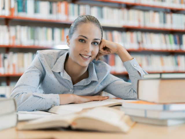 Smiling female student at the library, she is sitting at desk and studying, education and self improvement concept