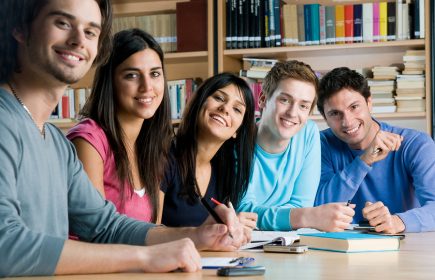 Happy group of young students studying together in a college library and looking at camera smiling